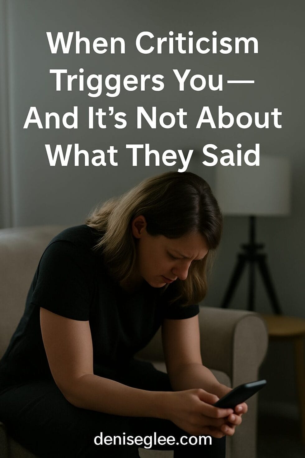 White woman in her 30s sitting on the edge of a couch, holding a phone with hunched shoulders and a troubled expression—visibly triggered by digital feedback
