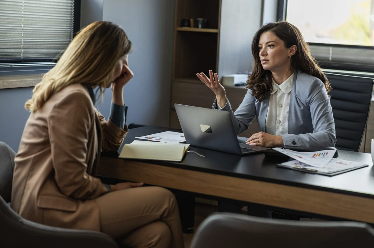 upset woman talking with another woman behind a desk with an open laptop