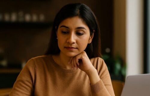 South Asian woman reflecting while writing in notebook with laptop nearby