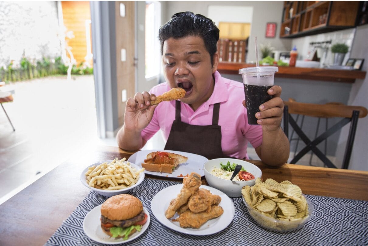 man sitting alone with tons of food and fried chicken near his mouth