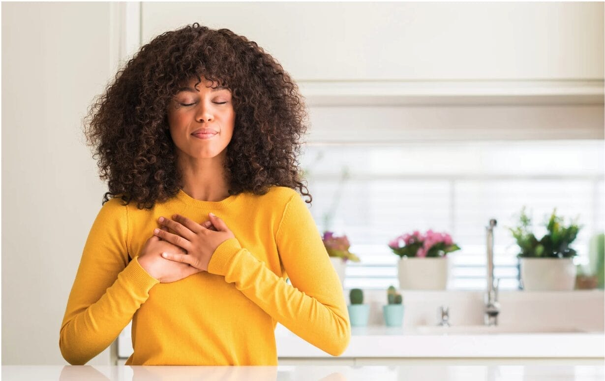 Black woman with curly hair in a yellow sweater, eyes closed and hands over her heart, standing peacefully in a kitchen with sunlight and flowers behind her