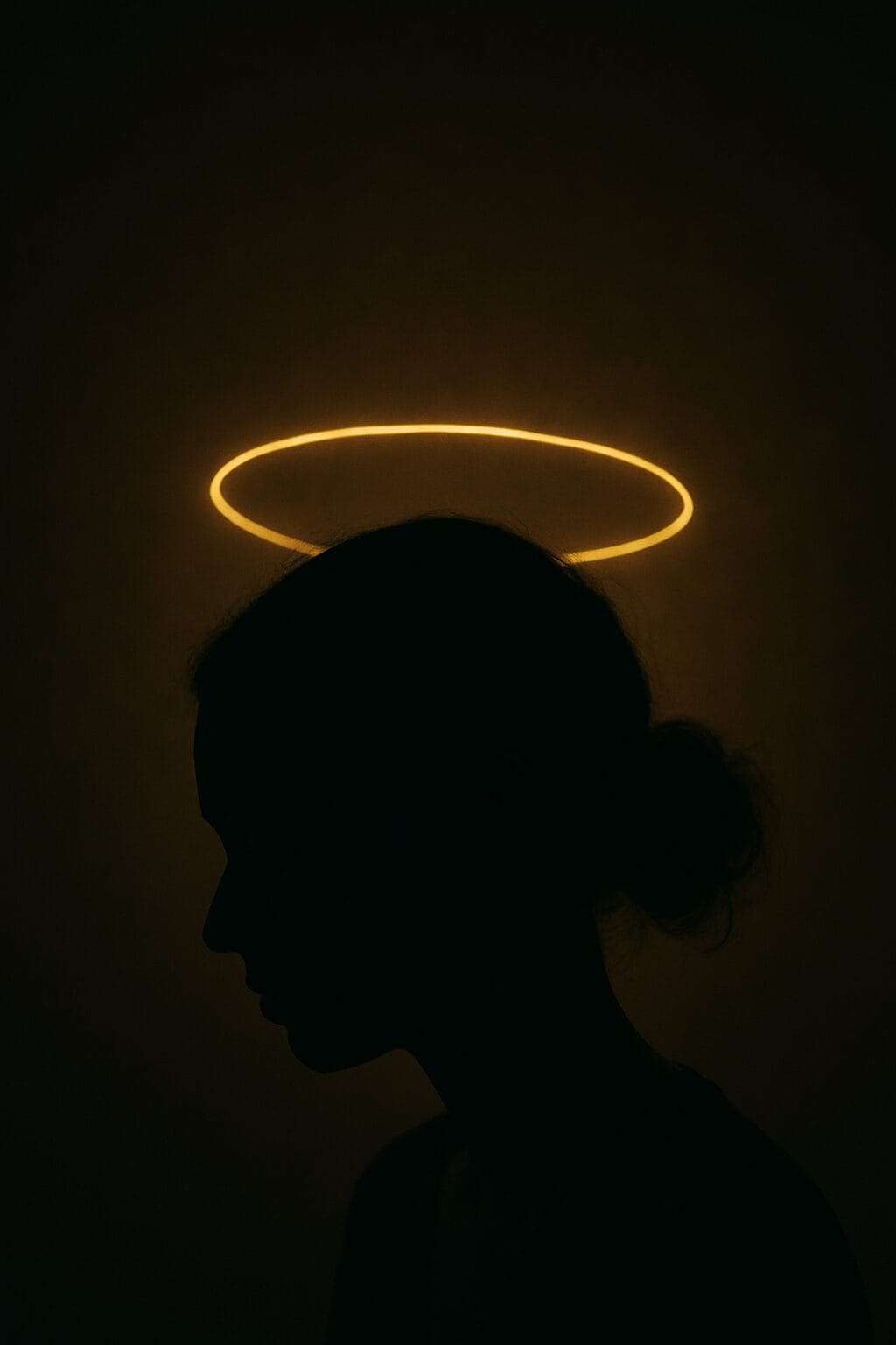 A digital photograph captures the dark silhouette of a woman’s head in profile, with a soft golden halo floating above, symbolizing the unseen emotional burden she carries.