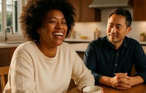 Mid-40s African American woman sitting at a luxury kitchen table with her Asian husband, both smiling softly in a quiet moment of connection and reflection